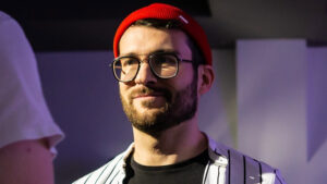 Porträtfoto
ein erwachsener Mann ist von den Schultern aufwärts zu sehen.
Er hat einen braunen Vollbart.
Er trägt eine rote Mütze, eine schwarz gerahmte Brille und ein schwarzes T-Shirt mit einem schwarz-weiß gestreiften Hemd darüber.
Der Hintergrund ist unscharf.